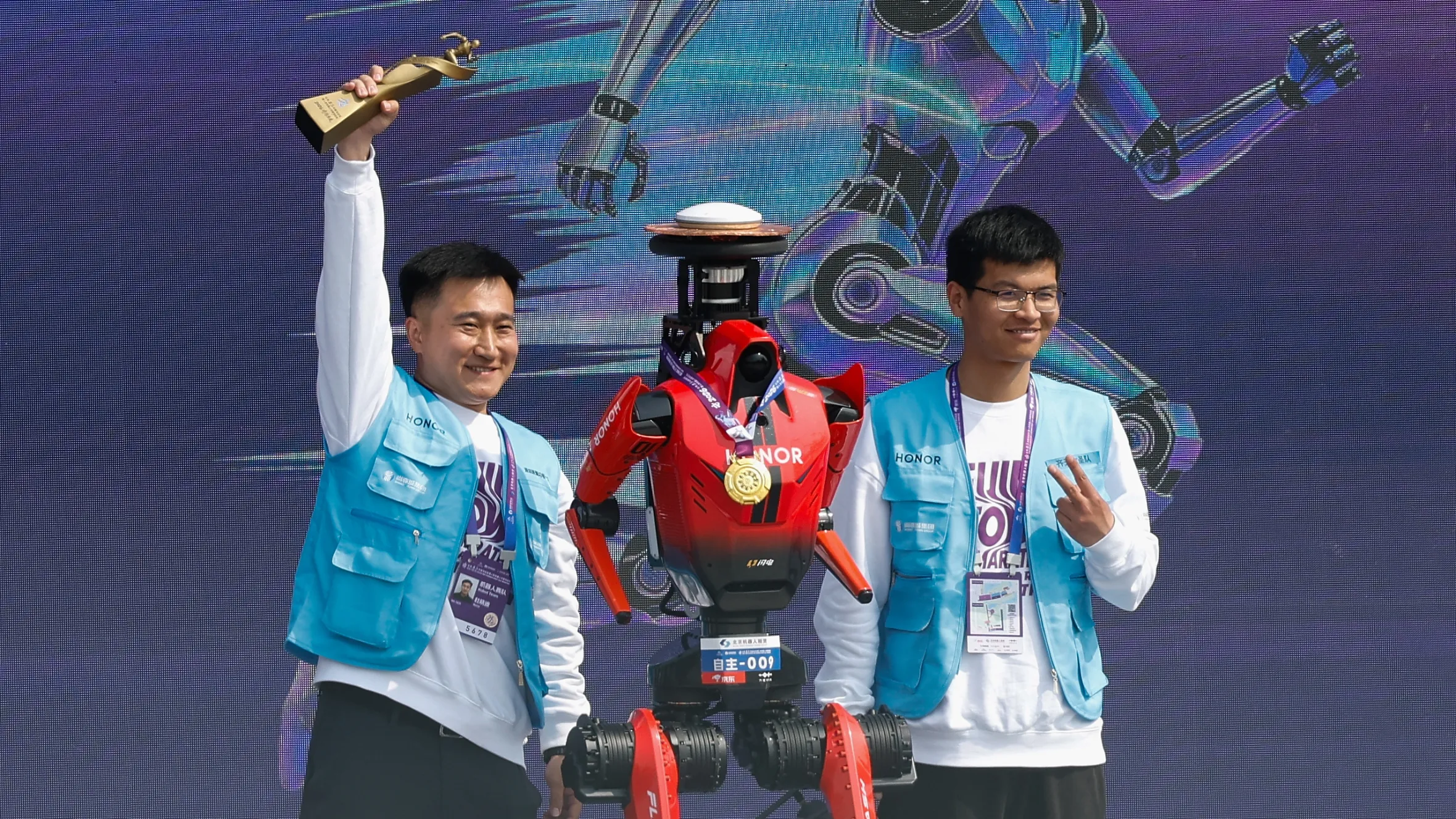 Two men and a red humanoid robot with a medal stand on a stage with an abstract blue background showing a running robot. One man holds a golden trophy above his head.