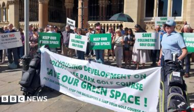 Lots of people stand in front of a stone building. They are holding green and white placards with "Keep our space" and "Save Collingtree Golf Club" written on them. There is also a large white banner bookended by two golf club bags with "Housing development on Collingtree Park Golf Course. Save our green space," written on it. It is a sunny day.