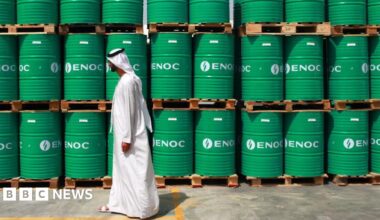 A man wearing a long, white dishdasha walks past stacks of bright green barrels bearing "ENOC" branding,