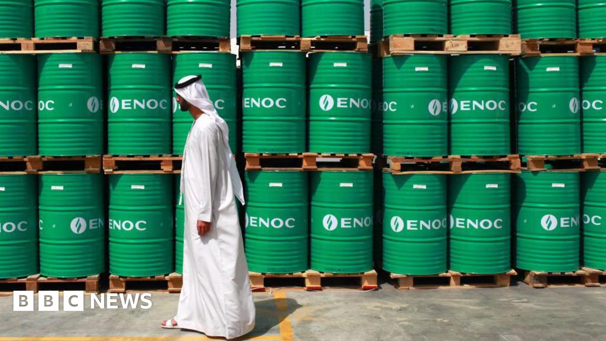 A man wearing a long, white dishdasha walks past stacks of bright green barrels bearing "ENOC" branding,