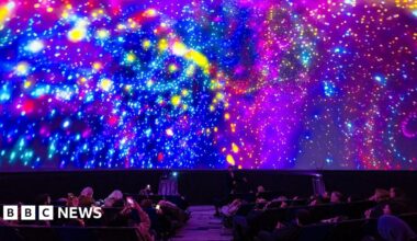 A large group of people recline in seats inside a dark planetarium-style dome while a vivid, colourful projection fills the entire curved ceiling. The display shows swirling patterns of bright neon colours - pinks, blues, yellows, and purples - resembling galaxies, stars, and abstract light bursts. Several audience members hold up phones to capture the immersive light show.