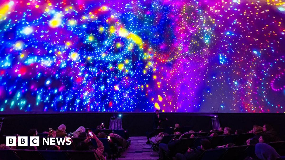 A large group of people recline in seats inside a dark planetarium-style dome while a vivid, colourful projection fills the entire curved ceiling. The display shows swirling patterns of bright neon colours - pinks, blues, yellows, and purples - resembling galaxies, stars, and abstract light bursts. Several audience members hold up phones to capture the immersive light show.