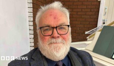 Alan Gregory, piano teacher, sits at a white piano in a dark three piece grey and navy suit. He has grey hair, a grey beard, blue eyes and wears glasses.
