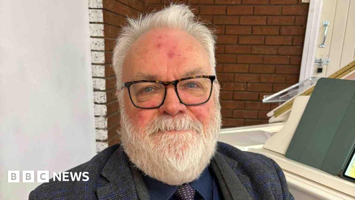 Alan Gregory, piano teacher, sits at a white piano in a dark three piece grey and navy suit. He has grey hair, a grey beard, blue eyes and wears glasses.