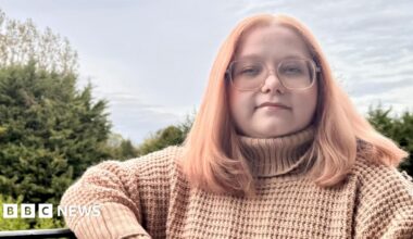 Beatrice Jones sitting on a bench outside. She has pink hair, glasses, and is wearing a pink jumper.
