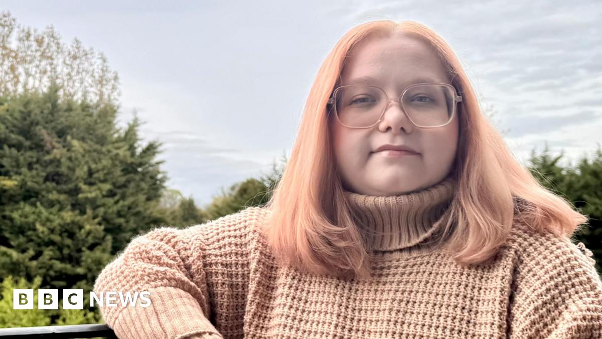 Beatrice Jones sitting on a bench outside. She has pink hair, glasses, and is wearing a pink jumper.