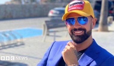 A man tanned smiling at the camera wearing aviator sunglasses a yellow Dsquared2 baseball cap and electric blue jacket and black T-shirt. He has a trimmed black beard. He is sat near a swimming pool which is out of focus behind him.