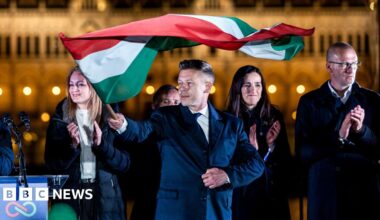 Magyar pictured in the dark holding the flag of Hungary and looking towards left of frame in front of other party members clapping with parliament in background as they celebrate after winning the election.