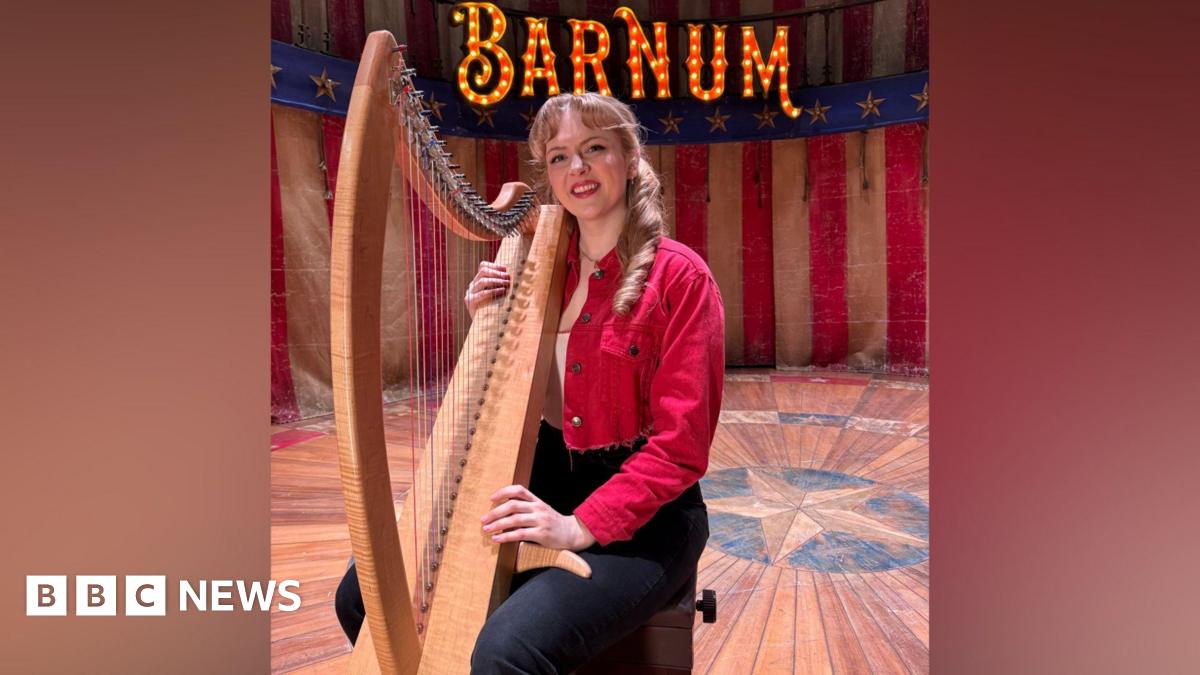 A young woman with dark blonde hair is sitting on a stool on a theatre stage with an American flag type background. She s wearing a red denim cropped jacket, white t-shirt and black jeans and is holding a large light brown harp with colourful strings. On the stage backdrop is the word "Barnum" in large letters lit up by bulbs