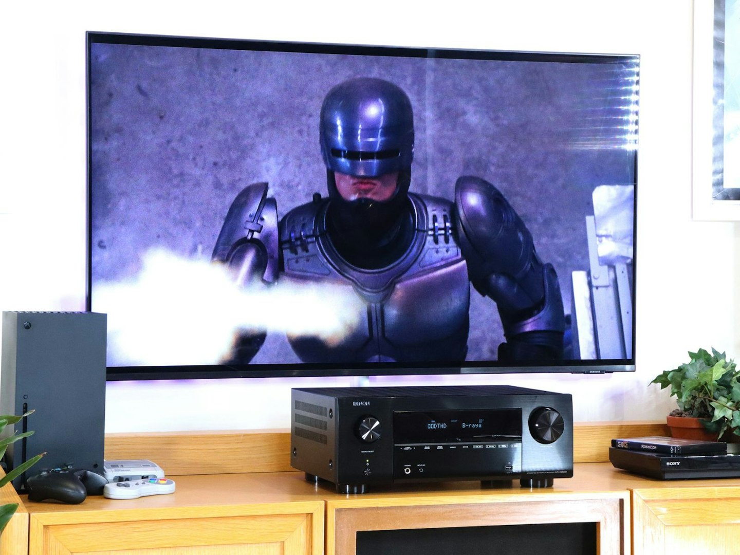 Denon AVR-X2800H Home Cinema Amplifier on a stand with Xbox and blu-ray player in front of a TV showing robocop