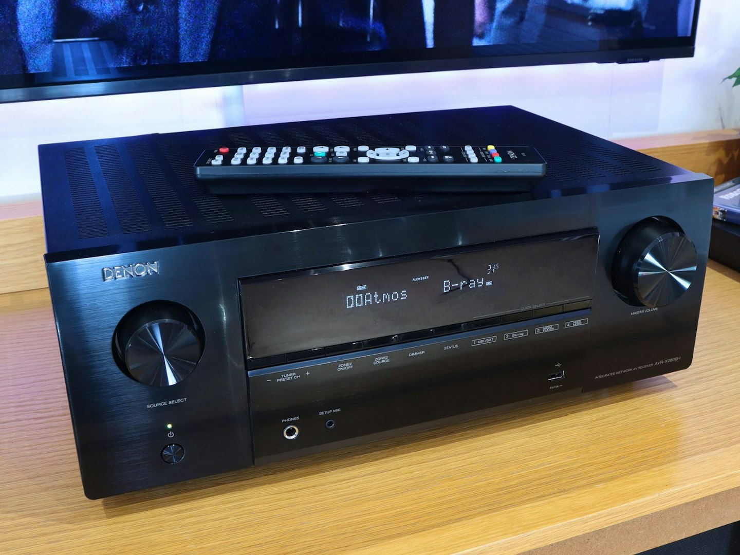 Denon AVR-X2800H Home Cinema Amplifier on a stand in front of a TV with remote