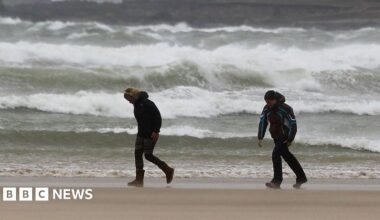 Two people bend into the wind as they walk along a beach with crashing waves behind them
