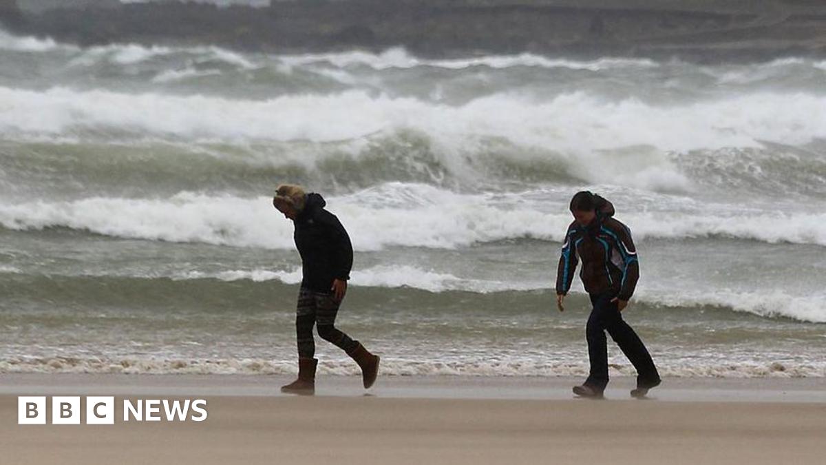 Two people bend into the wind as they walk along a beach with crashing waves behind them