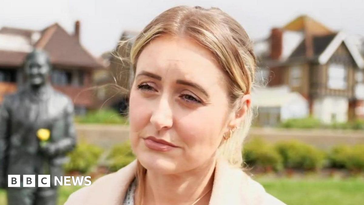 A blonde woman standing, looking past the camera. There is a metal statue over her right shoulder holding a yellow flower.