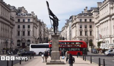 A grey statue of a man wearing a suit, marching forward, carrying a flag that covers his face