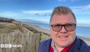 Councillor Scott Dickinson. He has short brown hair and is wearing glasses. He is standing in front of the beach of Druridge Bay.
