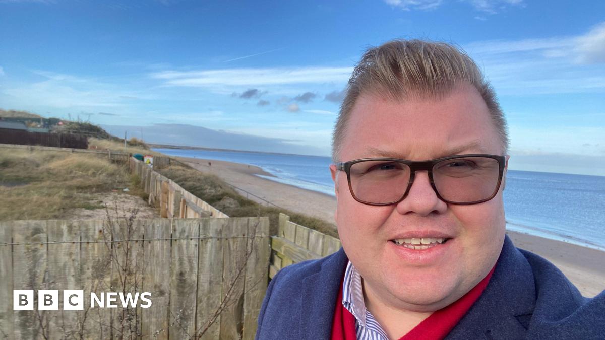 Councillor Scott Dickinson. He has short brown hair and is wearing glasses. He is standing in front of the beach of Druridge Bay.