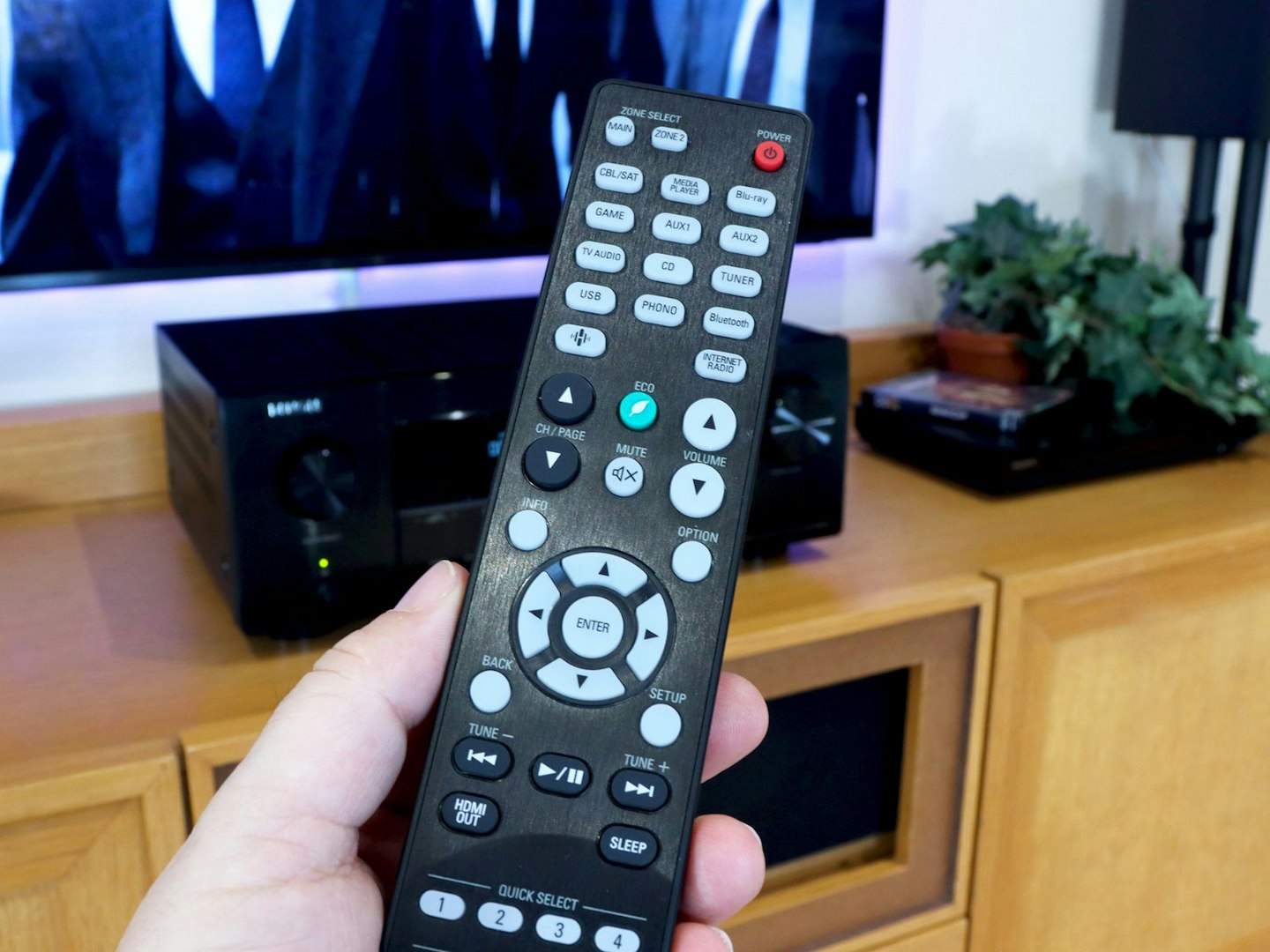 Denon AVR-X2800H Home Cinema Amplifier on a stand in front of a TV and someone holding the remote