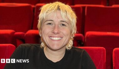 Maisie has short, shaggy blond hair and sits smiling in a theatre auditorium, leaning forward with hands clasped. She is wearing a black T‑shirt and sits among rows of bright red seats that fill the background.
