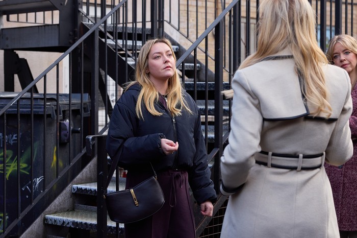 Megan sits on a staircase, confronted by Eva and Leanne in Coronation Street