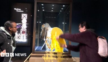 Two people stood next to the display case holding the Crown Jewels in the Tower of London, one is stood looking on while another is throwing yellow custard at the glass.