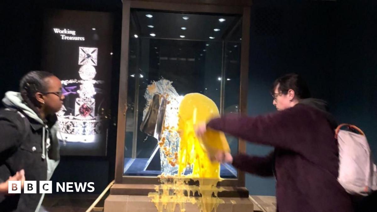 Two people stood next to the display case holding the Crown Jewels in the Tower of London, one is stood looking on while another is throwing yellow custard at the glass.