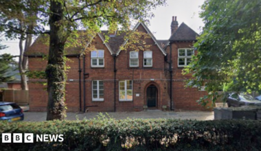 A red-brick building stands behind trees and bushes. The building has two apex roof sections and six large white-framed windows. It has a door arch for an entrance and a paved area out the front used as a car park.