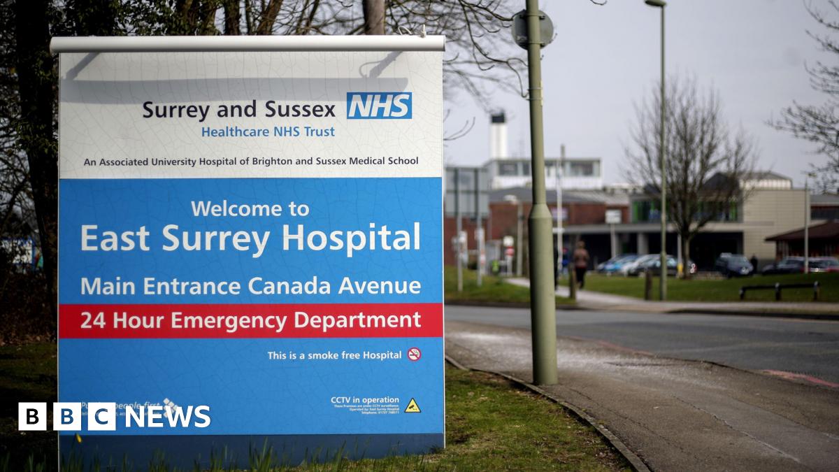 A large blue and white sign at the entrance to East Surrey Hospital shows directions to the main entrance and 24‑hour emergency department. The sign stands beside a road with streetlights, trees and hospital buildings visible in the background.