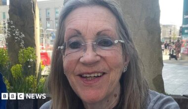 A selfie of Margaret Colling sitting outside in front of two tall decorative stones and planters. She is smiling and has long, straight greying hair and rim-less glasses