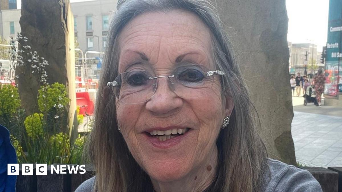 A selfie of Margaret Colling sitting outside in front of two tall decorative stones and planters. She is smiling and has long, straight greying hair and rim-less glasses