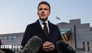 A woman and a man in black jackets look out over Tehran's skyline with a tall tower in the distance. They have their backs to the camera, she has long brown hair tied in a pony tail and her right hand rests on his left shoulder.
