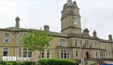 A long large Victorian building takes up most of the image with steps leading up to the entrance and a ramp to one side. A large clock tower sits in the centre of the building and there are several chimneys
