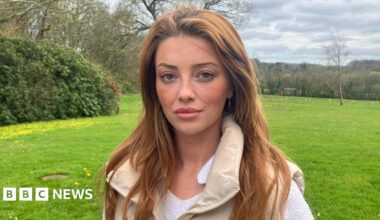 A woman with strawberry blonde hair stares at the camera. She's stood at a gate looking over a green field behind her, with hedges and trees in the background. The sky has grey clouds with a hint of blue skies in the distance. Lorna is wearing a white top, with a cream gillet over the top.