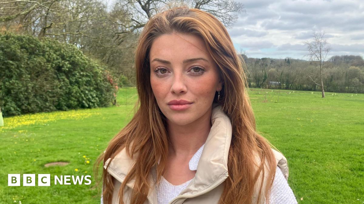 A woman with strawberry blonde hair stares at the camera. She's stood at a gate looking over a green field behind her, with hedges and trees in the background. The sky has grey clouds with a hint of blue skies in the distance. Lorna is wearing a white top, with a cream gillet over the top.
