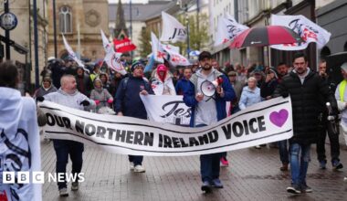 A group of people marching down a street during a rally. They are holding a white banner which says: "Our Northern Ireland Voice". It is during the day. Many of the people are wearing coats and hats and some are holding flags and some umbrellas.
