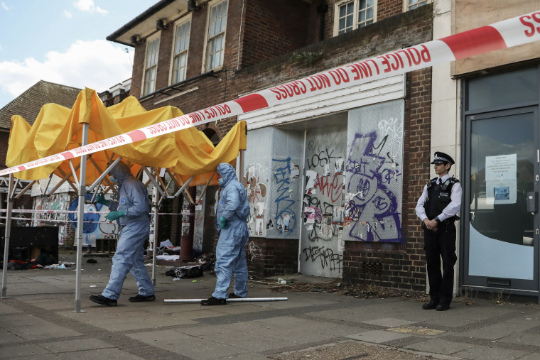 A crime scene with red and white striped tape surrounding it and two people in blue haz-mat suits.