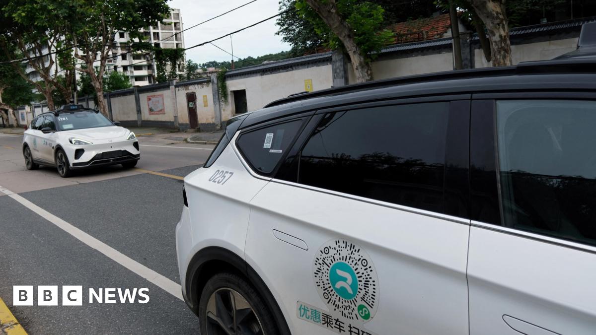 Two white driverless cars with black hoods are stationary on the road, one behind the other