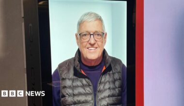 Pam stands smiling broadly next to the hologram version of Bill which is a picture of him smiling on a large screen