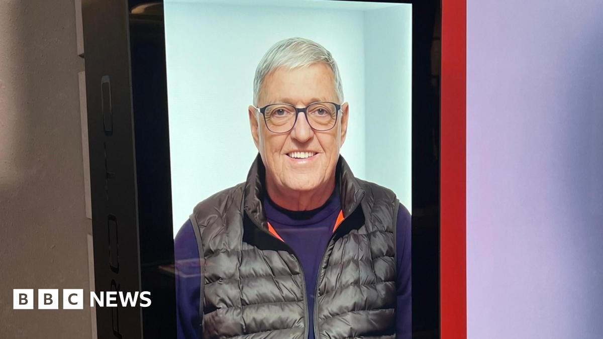 Pam stands smiling broadly next to the hologram version of Bill which is a picture of him smiling on a large screen