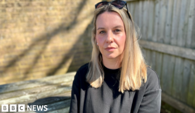 Zoe Trafford has long brown hair and wears a grey blazer and white top underneath. She is sitting against a background of wooden blinds against windows. The leaves of an indoor plant are also there.
