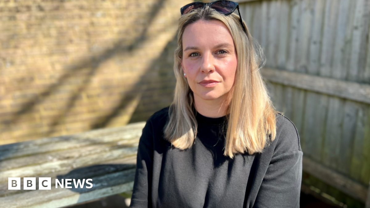 Zoe Trafford has long brown hair and wears a grey blazer and white top underneath. She is sitting against a background of wooden blinds against windows. The leaves of an indoor plant are also there.