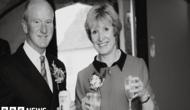 Photo of Tacey and David at a wedding. It is a black and white photo. David is wearing a suit and tie and holding a can or beer and a glass of beer in both hands.  Tacey is smiling to the camera holding wine glasses in both hands.