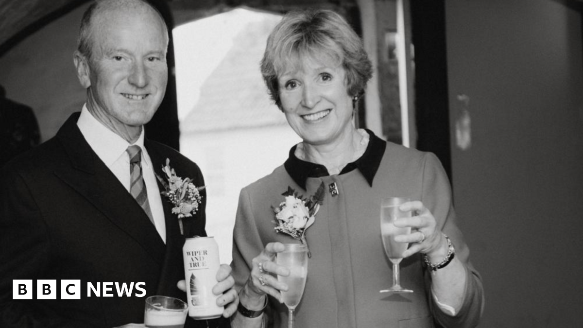 Photo of Tacey and David at a wedding. It is a black and white photo. David is wearing a suit and tie and holding a can or beer and a glass of beer in both hands.  Tacey is smiling to the camera holding wine glasses in both hands.