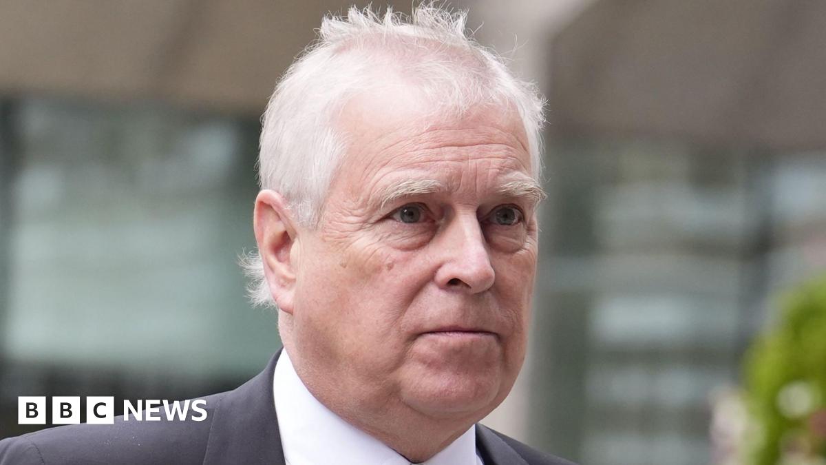 Andrew Mountbatten-Windsor, with short white hair in a black suit and tie, walks past a glass building