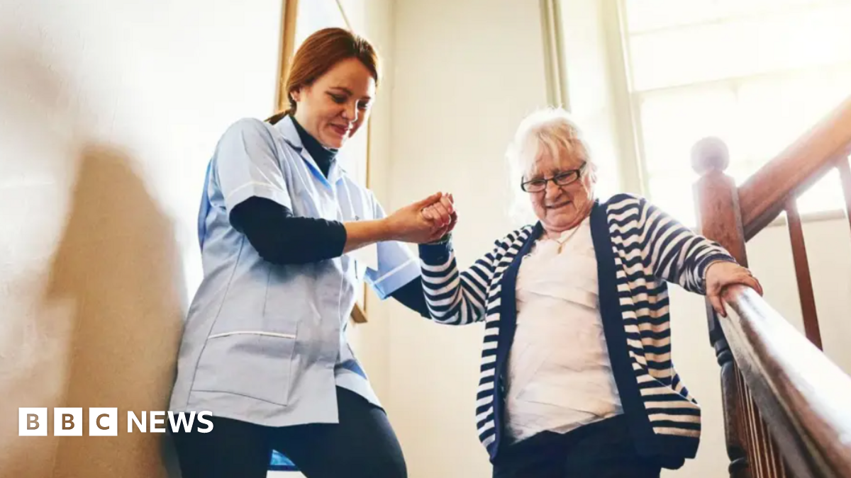 A care worker helping an elderly woman walk down a staircase. The carer, on the left, is wearing a light-blue jacket and dark trousers. She is smiling and holding the older woman's hand. The older woman has white hair, is wearing spectacles and has a look of concentration on her face as she takes the steps. She is holding the banister with her left hand.