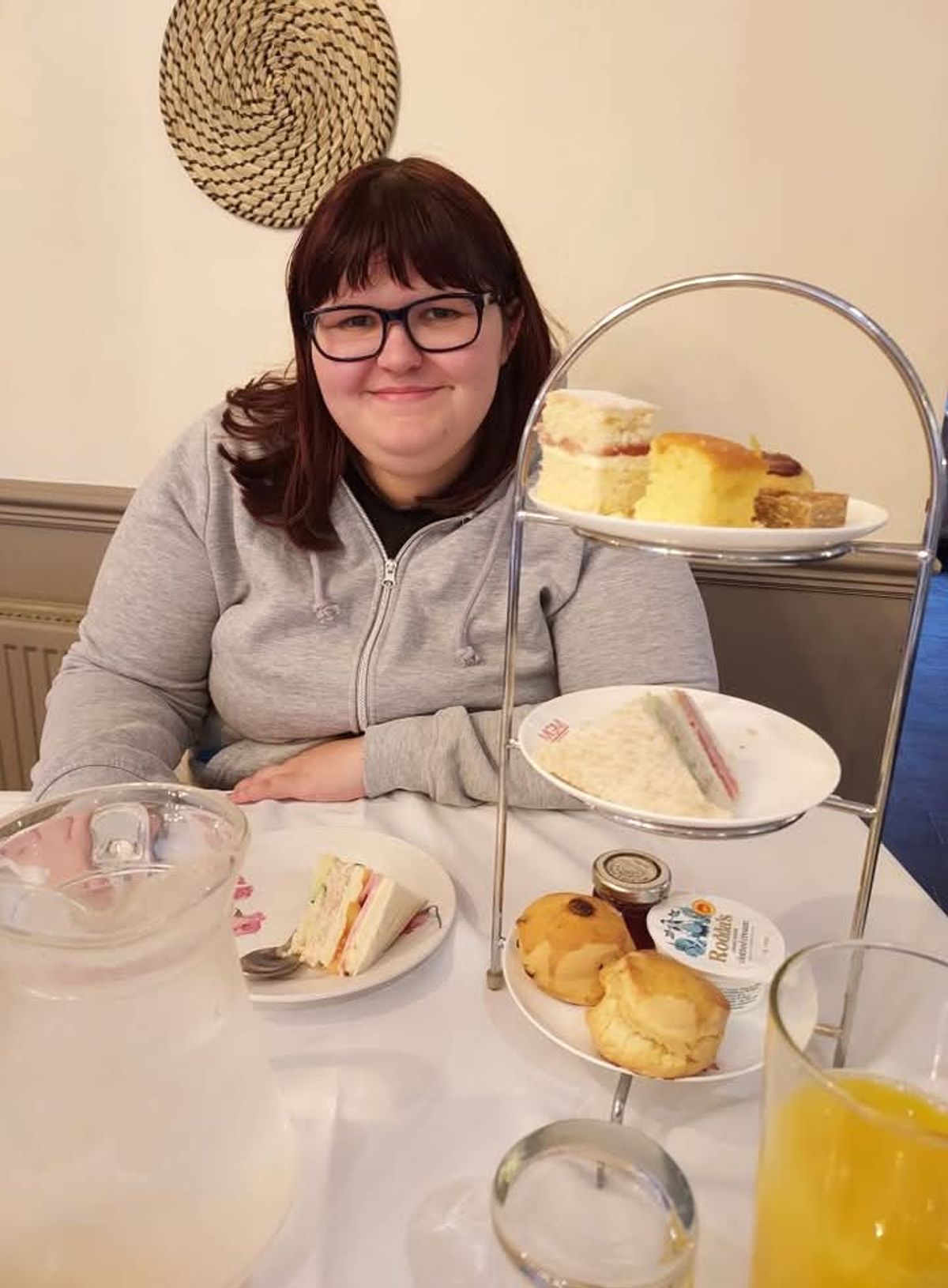 Woman with reddish hair smiling at camera, sitting behind dining table with scones and sandwiches as part of an afternoon tea