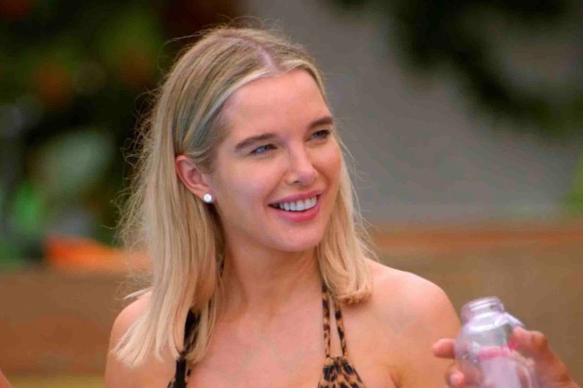 An individual with long, blonde hair wearing a black bikini top is smiling while standing in an outdoor setting, with a blurred background of greenery and a wooden structure.