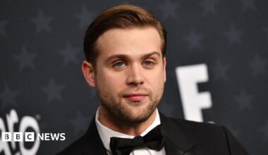 The headshot of Leo Woodall shows him wearing a white shirt with a black suit jacket and bowtie. He stands against a grey background with stars and has short brown hair and green-blue eyes.