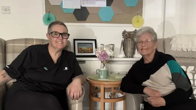 A woman with black glasses and short hair and an elderly woman with glasses and short grey hair. They are sat in arm chairs and smiling at the camera in front of them.