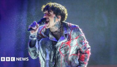 Bring Me The Horizon band on the Brit Awards red carpet. Left to right as we look are Matt Kean, Matt Nicholls, Oli Sykes and Lee Malia. All four men are wearing black. Matt Nicholls has black sunglasses on and tattoos visible on his hands. Oli also has tattoos covering both hands and is wearing an almost see-through black netting style top.